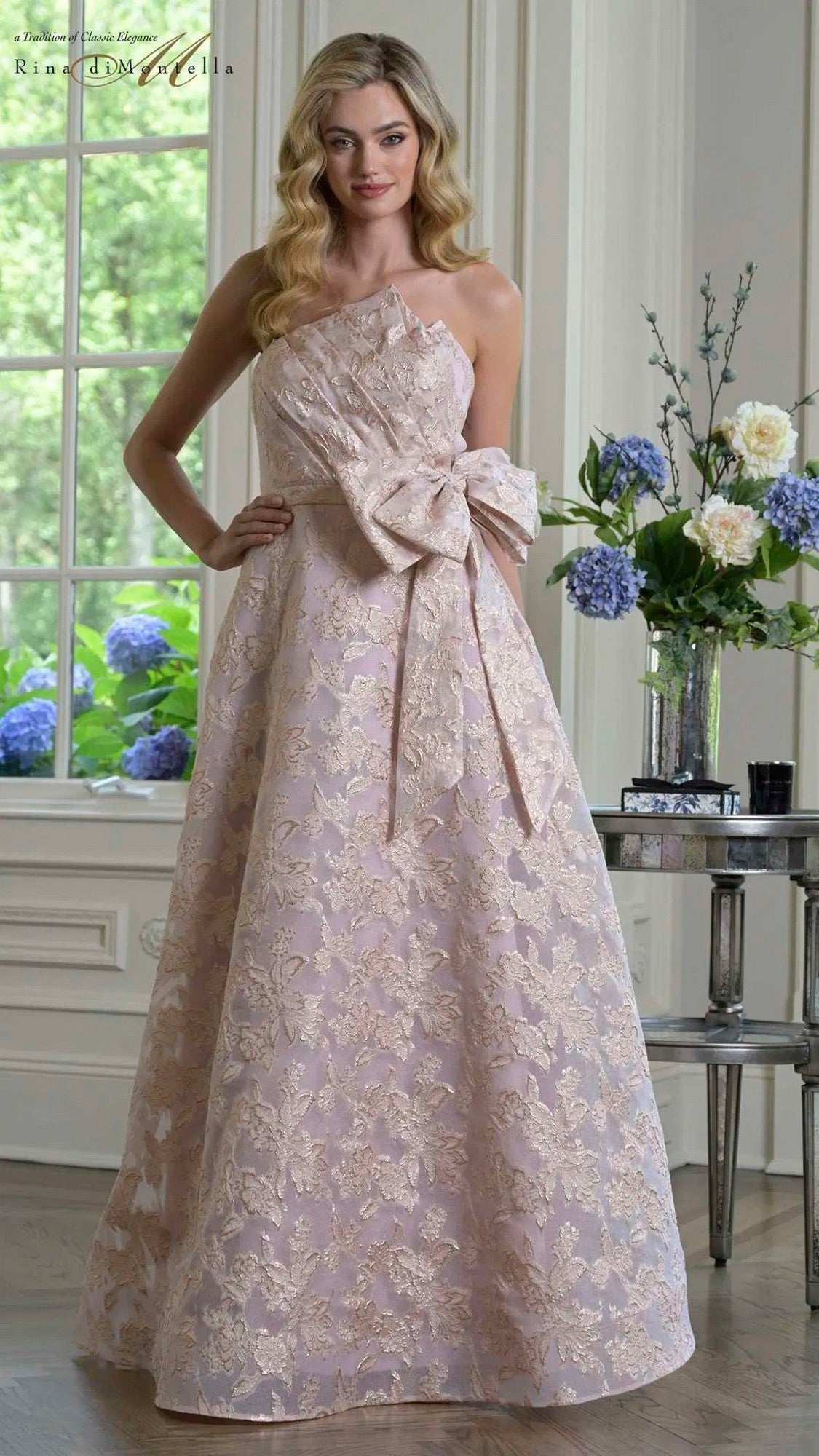 A woman stands indoors wearing the pink Long Formal Dress: Rina Di Montella RD3427, a floor-length gown with lace, an asymmetrical neckline, and a large bow at the waist. Blue and white flowers appear near a window in the background.